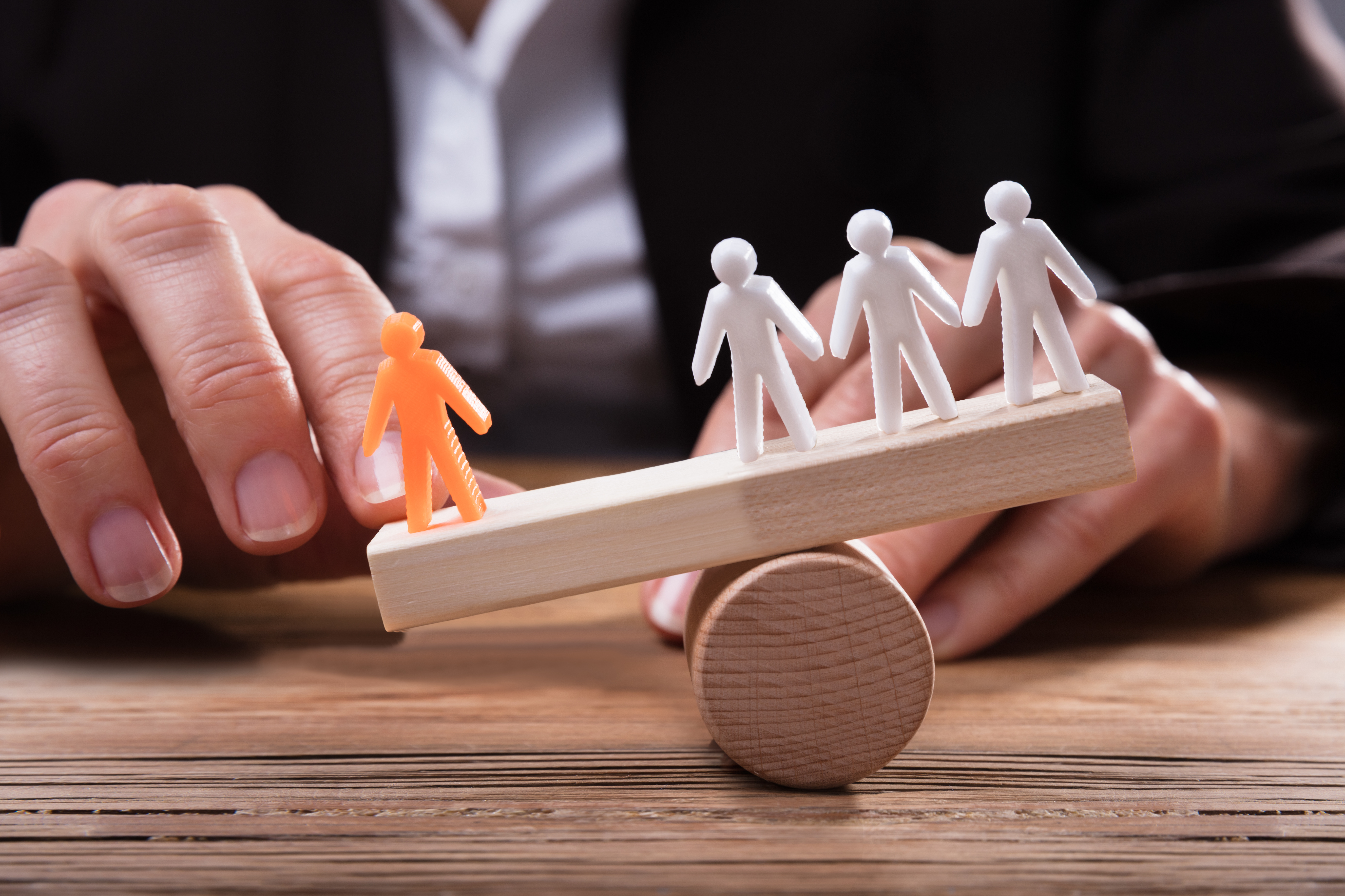 Close-up Of Businessperson Showing Orange Figure Against White Human Figures On Seesaw Over Wooden Desk
