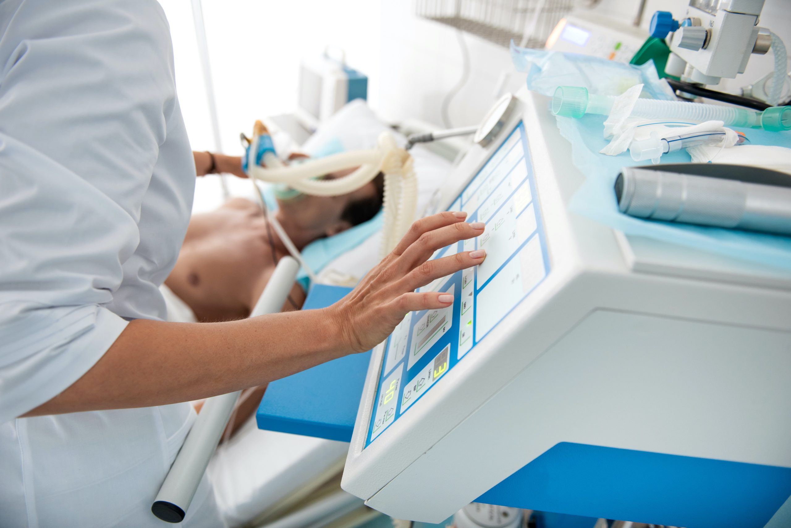 Close up of female hands touching monitor of mechanical ventilator. Middle aged man lying in hospital bed on blurred background