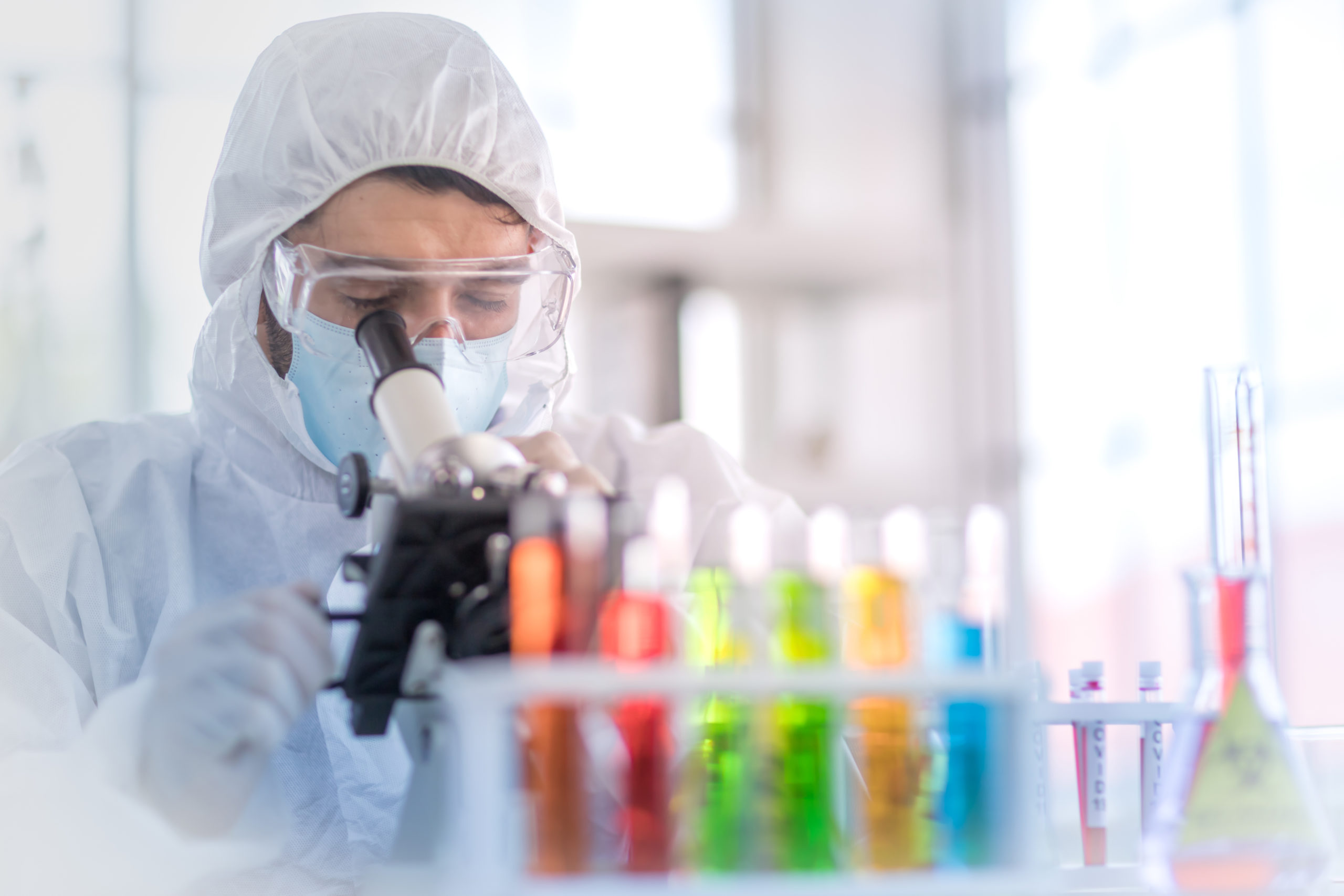 A male scientist wearing a mask and looking through the microscope in the laboratory, Concept, The scientists wear white protective clothing is experimenting with drug anti- Coronavirus