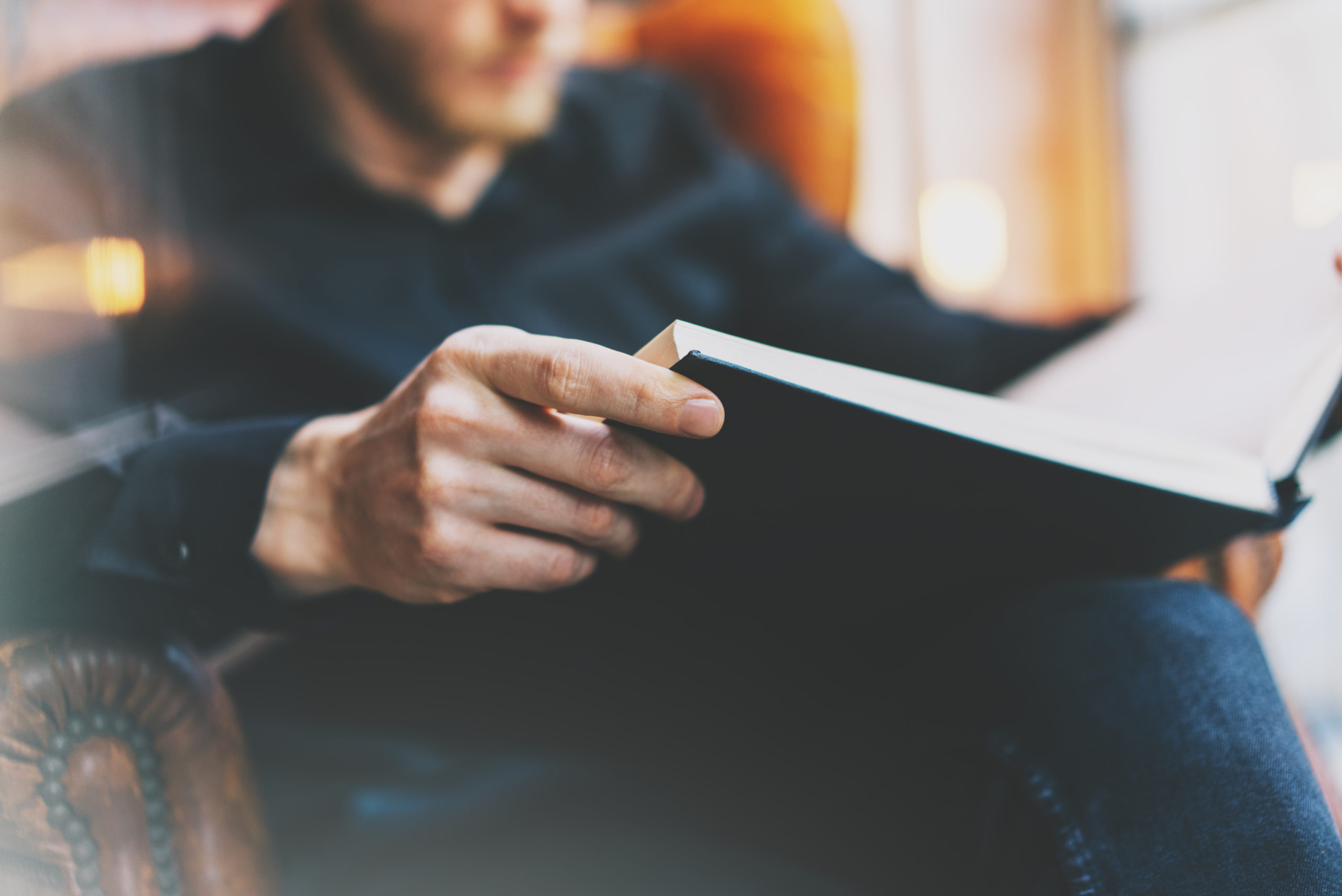 Portrait handsome bearded man wearing glasses black shirt.Man sitting in vintage chair university library, reading book and relaxing. Blurred background.Horizontal, film effect.