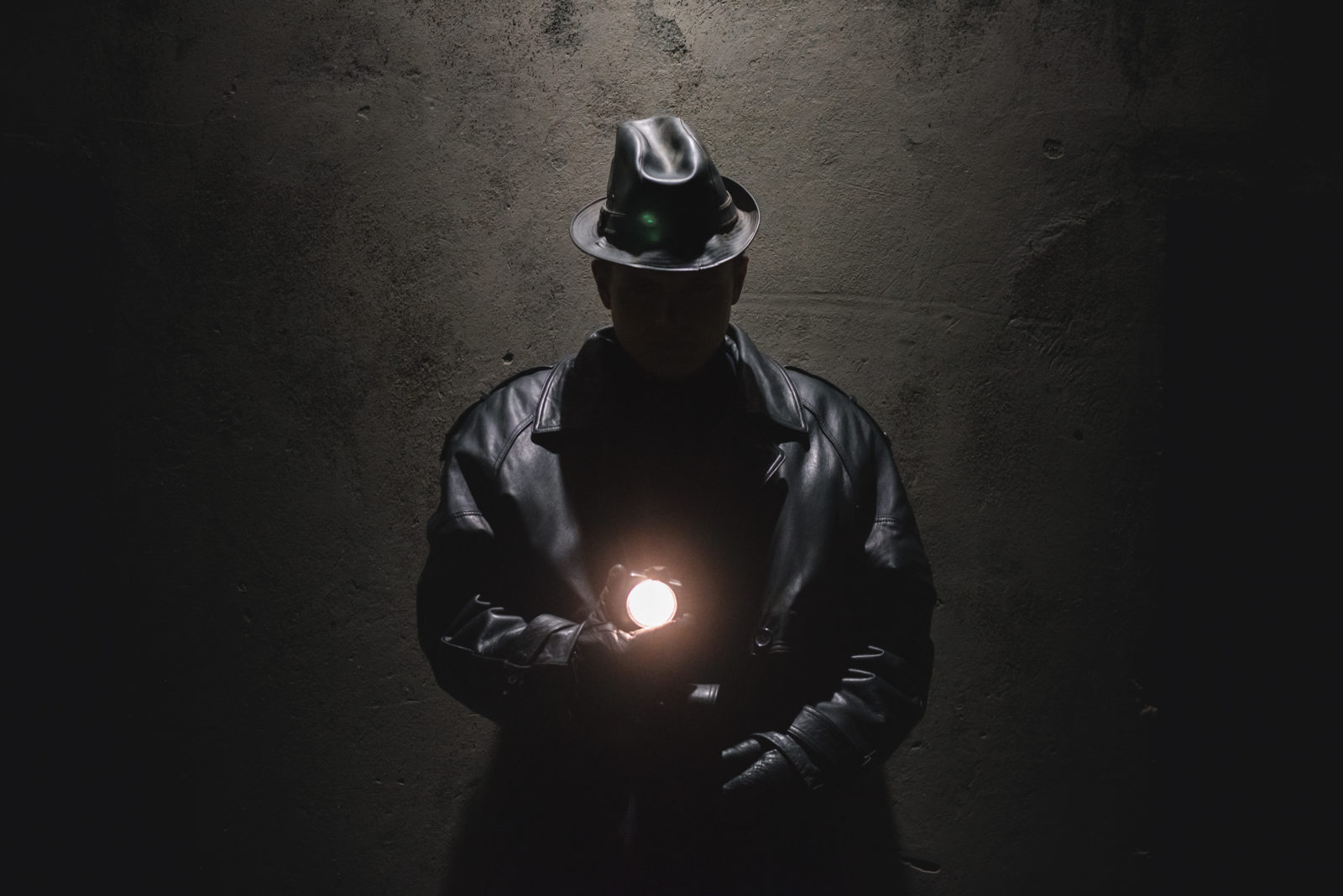 Detective agent in black coat and in leather hat is standing near the building wall at night.