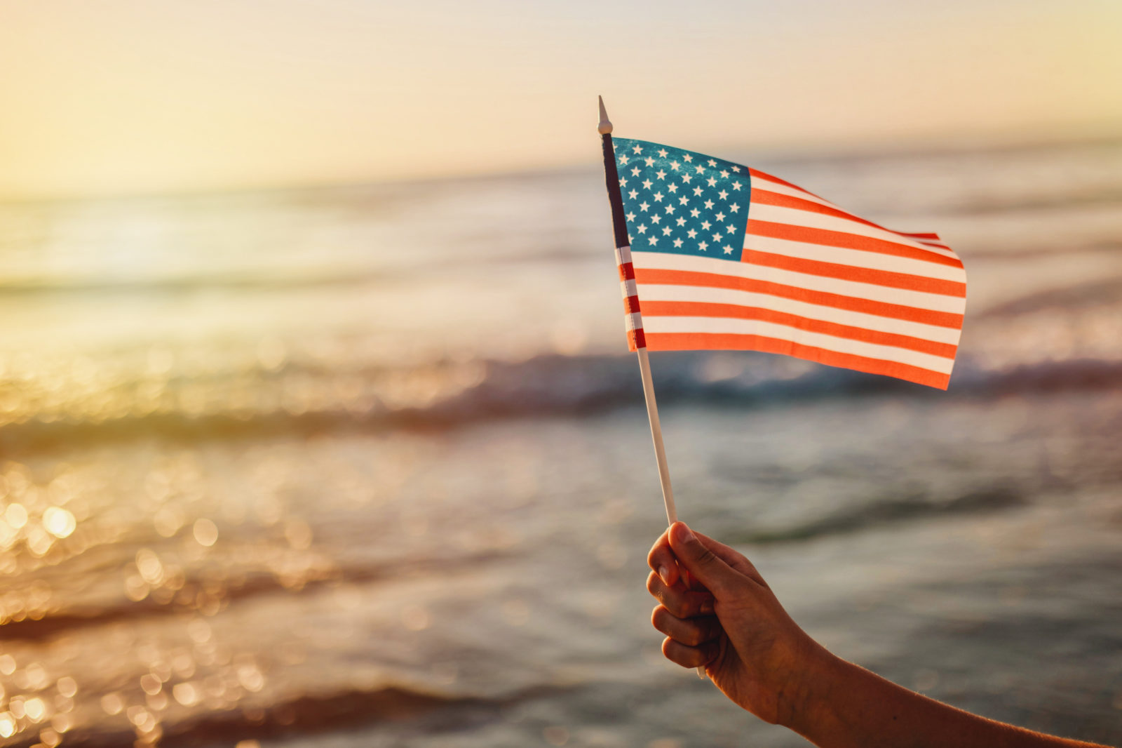 Close-up Of Hand Holding American Flag Against Sea During Sunset