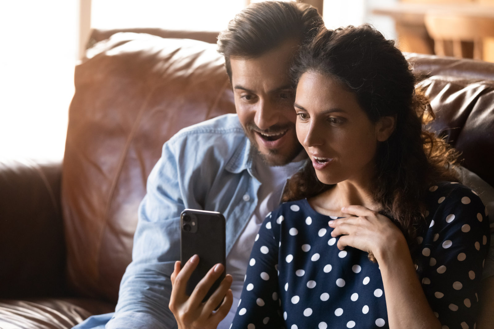 Shocked happy couple with smartphone reading message, getting surprising unbelievable good news from video call, using mobile phone together, staring at screen in amazement, sitting on couch at home