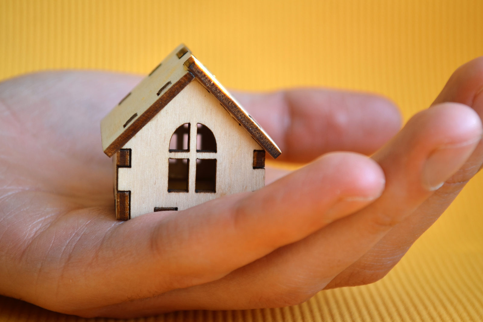 Wooden toy house model in man's hand on yellow background front view