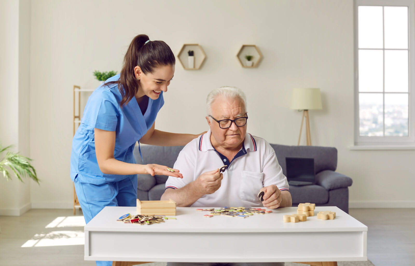 Friendly smiling nurse caregiver in geriatric clinic or retirement home helps senior man with Alzheimer's disease. Happy old man plays puzzles while sitting at table in cozy interior. Dementia concept