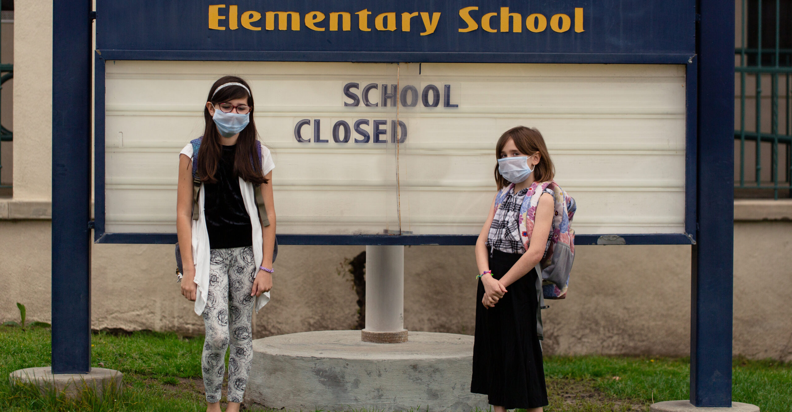 Two school aged girls are standing in front of their closed school with facemasks on during the outbreak of covid-19.