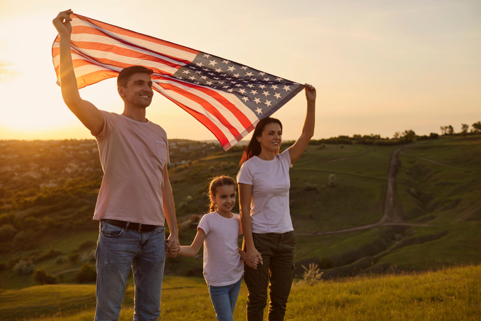 Young parents with their daughter holding American flag in countryside at sunset. Independence Day celebration