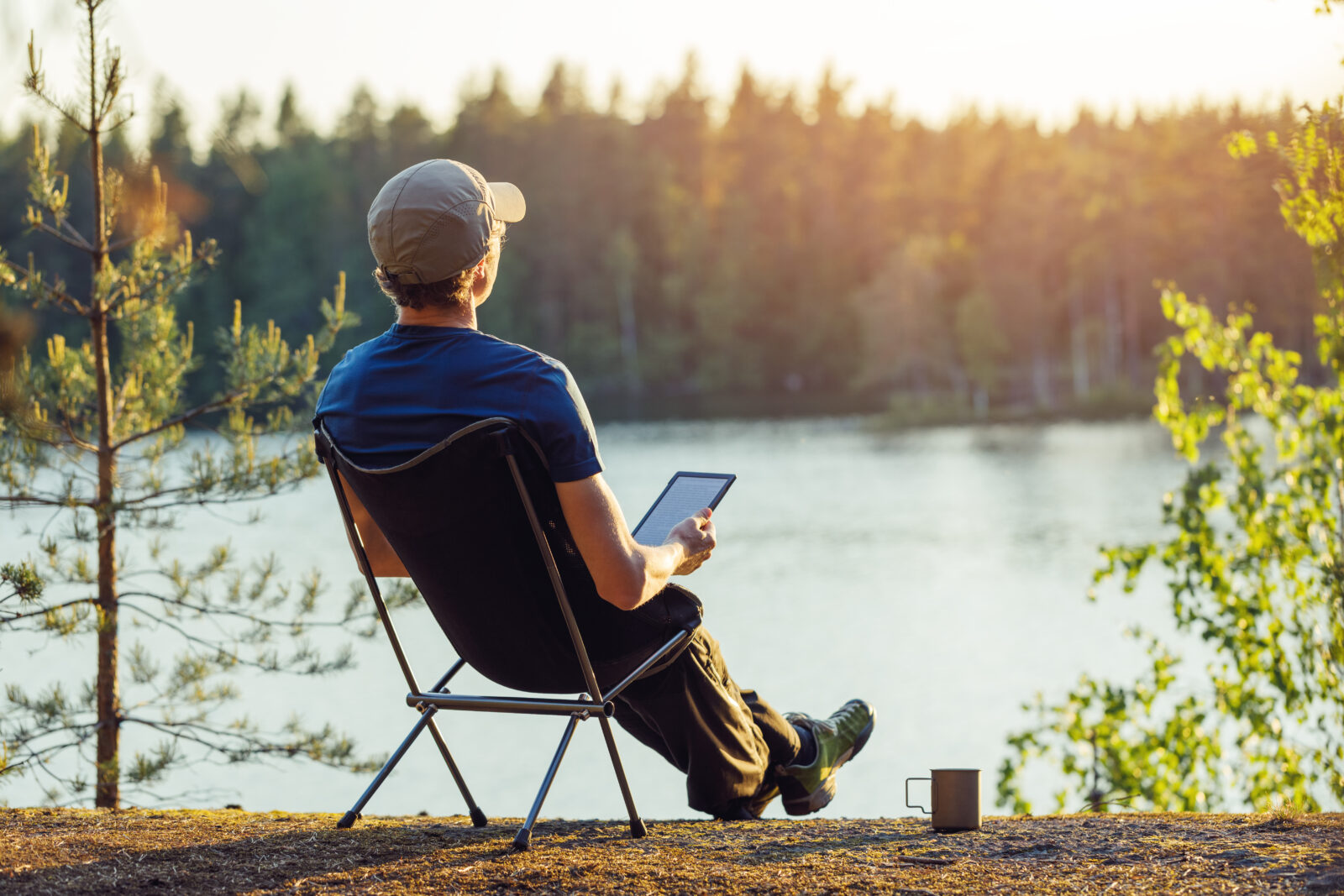 Man reading an e-book on the shore of a forest lake at sunset.