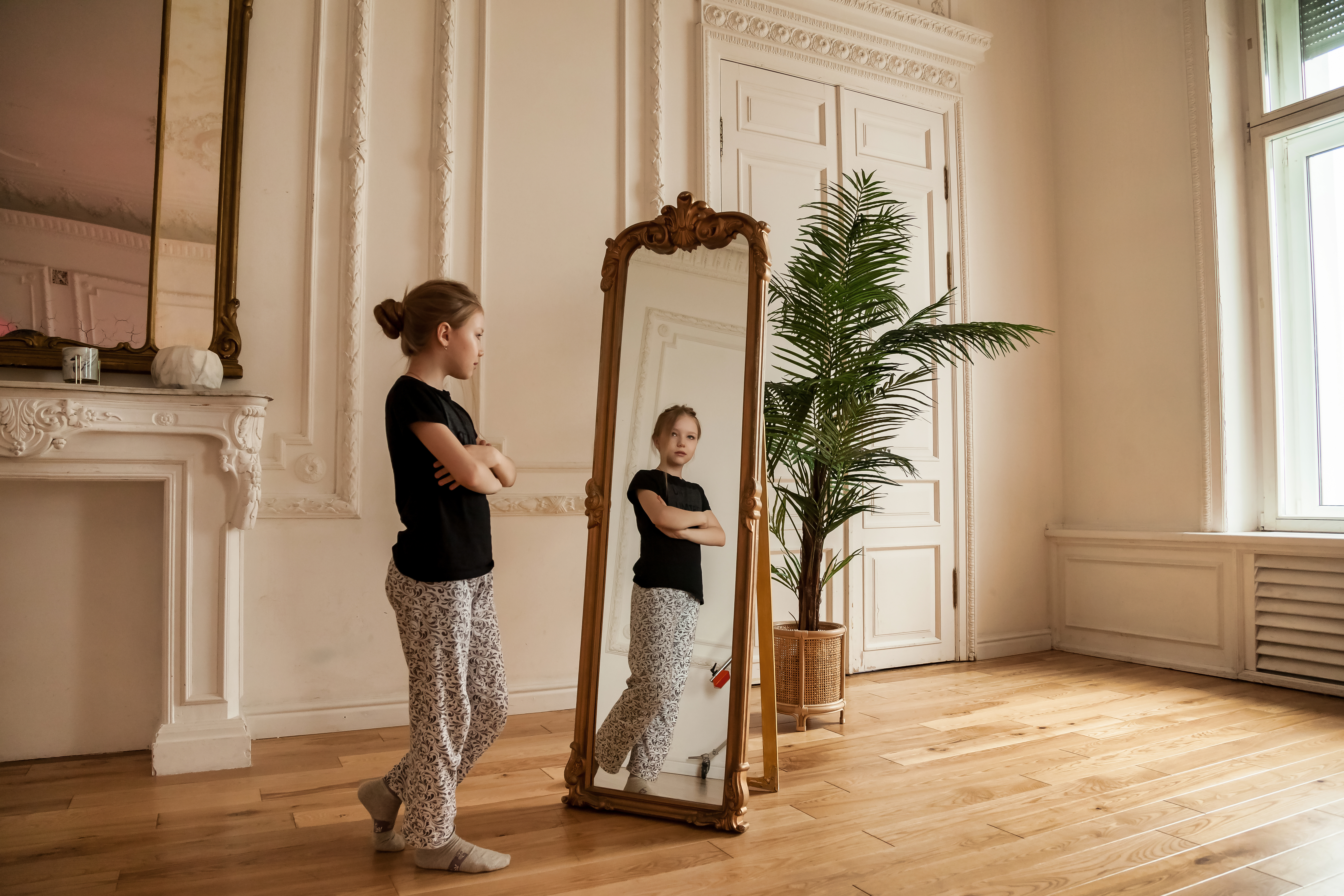 Alone cute teenager girl at mirror in living room at home, looking her. Sad lonely child stands and looks in his reflection in casual domestic clothes. Concept difficult childhood. Copy text space