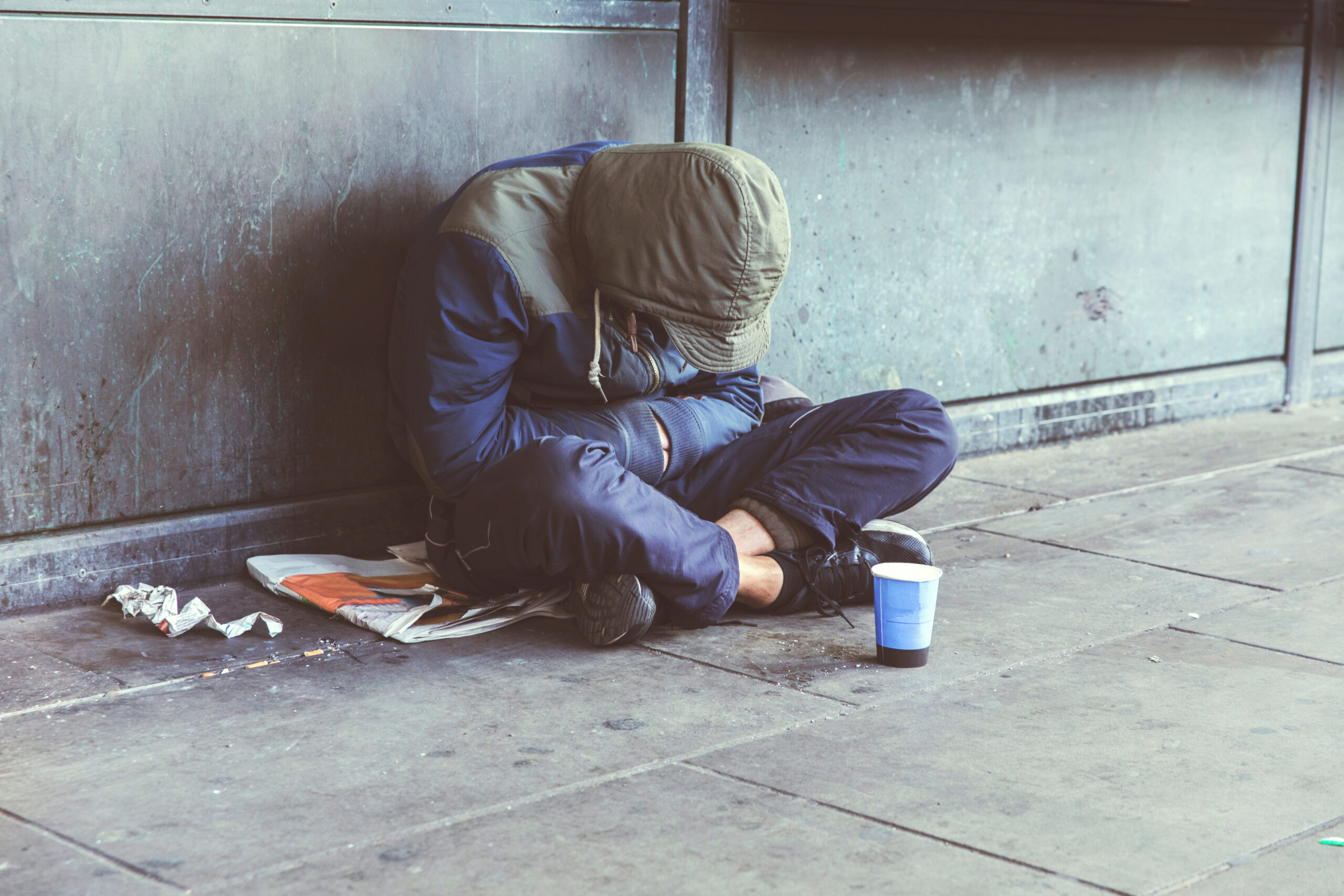 Homeless adult man sitting on the street in the shadow of the building and begging for help and money. Problems of big modern cities. Indifference of people. Social issues.