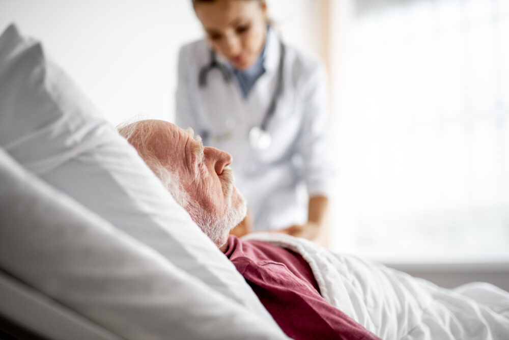 Side view portrait of bearded gentleman lying in bed. Young woman in white lab coat on blurred background