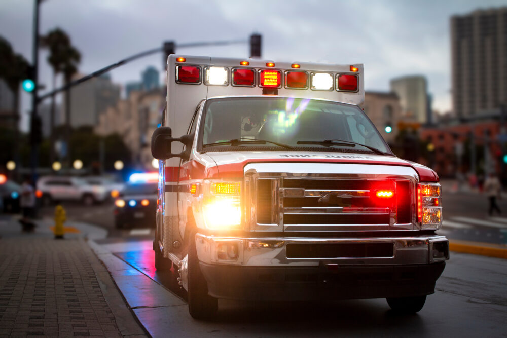 An ambulance with lights activated and a police car behind it in an urban environment