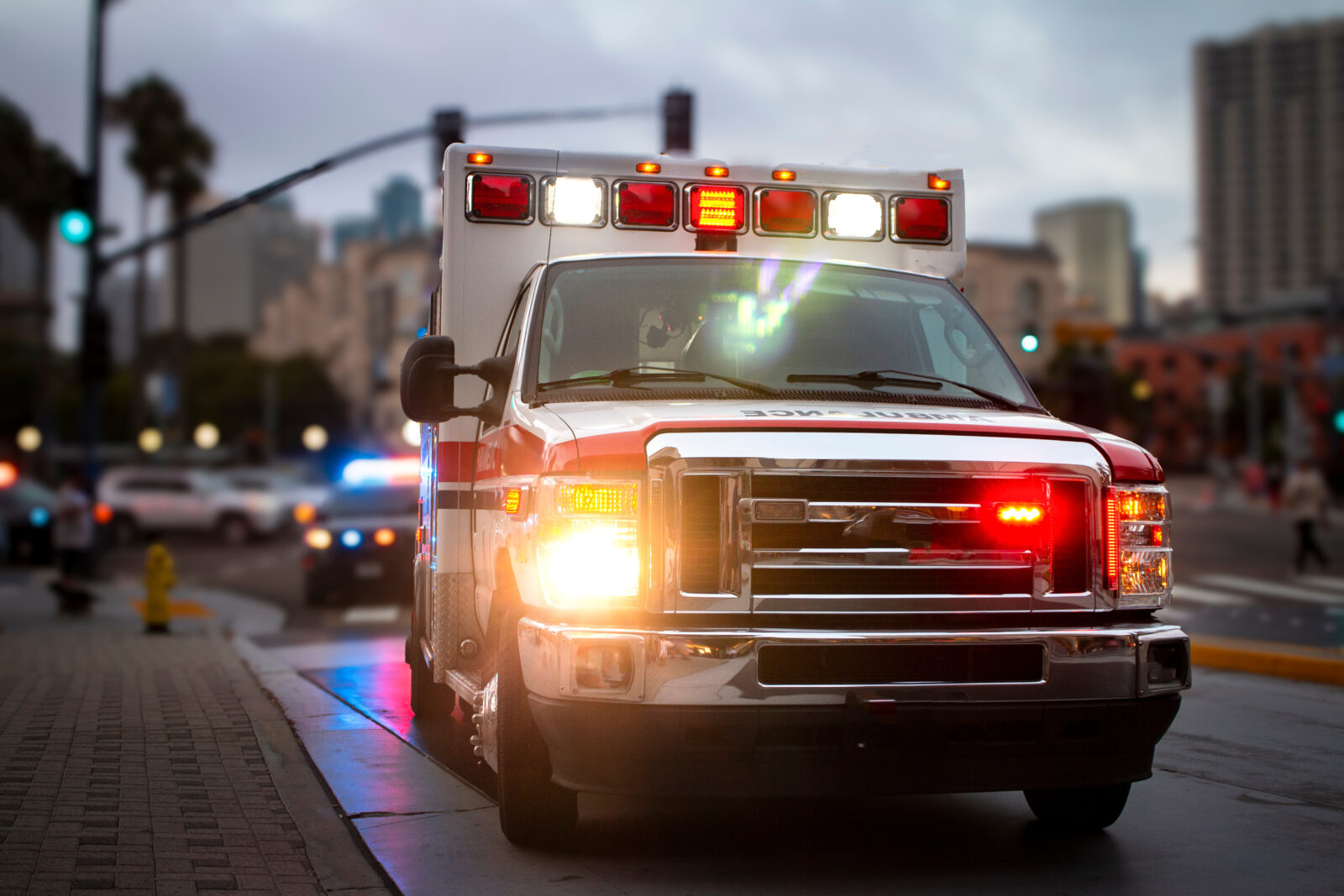 An ambulance with lights activated and a police car behind it in an urban environment