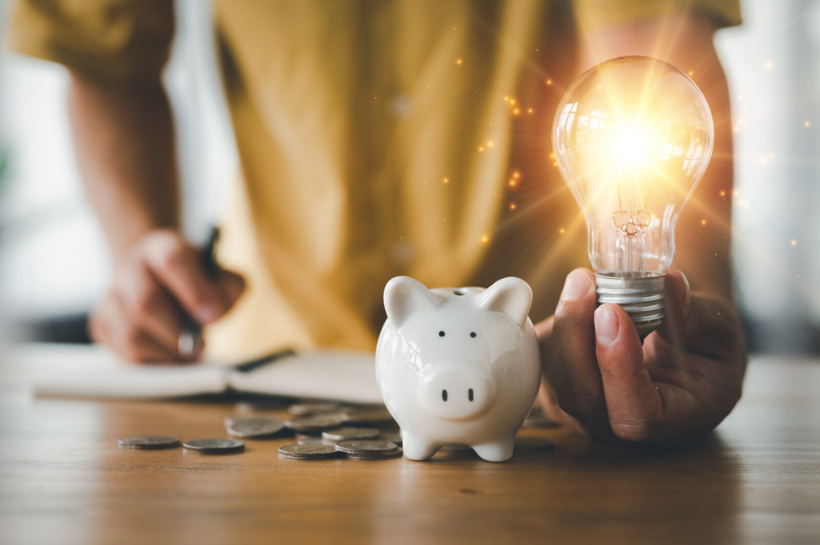 saving energy and money concept. idea for save or investment. businessman holding lightbulb beside piggy bank and coins stacking on desk with note book.