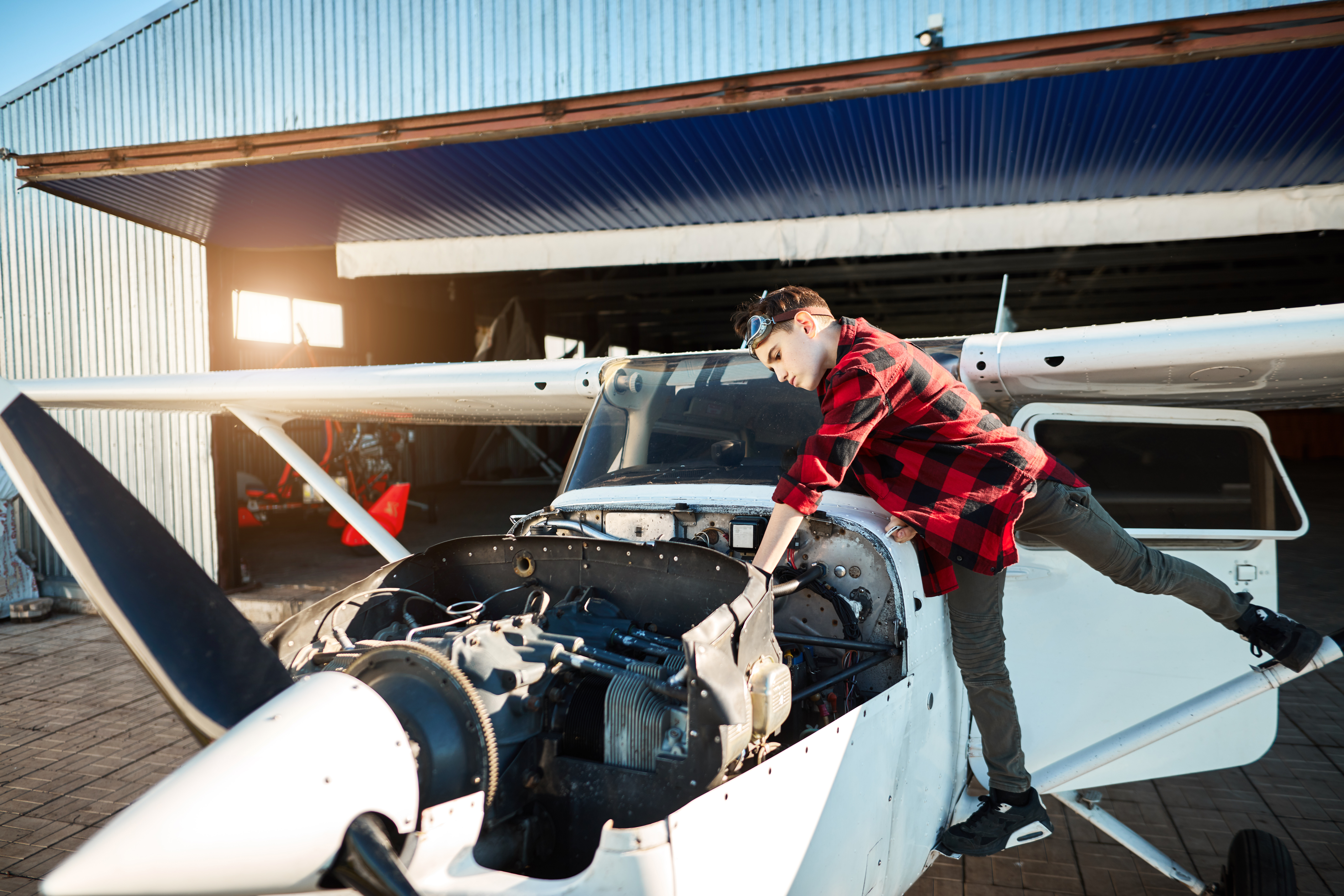 view of light private propeller airplane with opened motor cabinet stands outside hangar, teenager boy standing near, observing motor compartments, waiting his father to come.