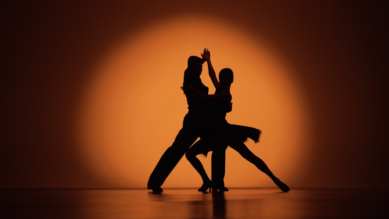 Couple of dancers approach each other and begin to dance Argentine tango. Elements of latin ballroom dance in studio with orange brown background. Dark silhouettes. Slow motion ready, 4K at 59.94fps.