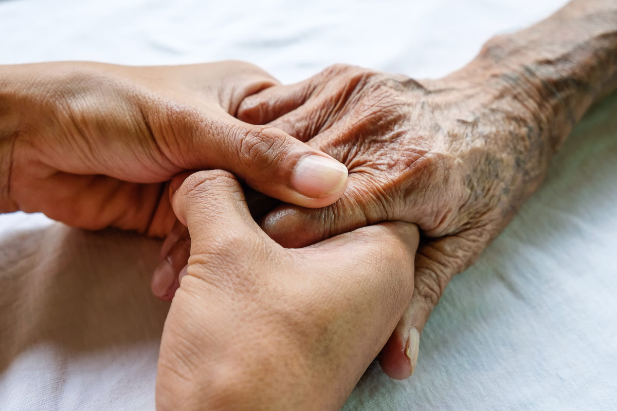 Hands of the old man and a young man on a white bed in a hospital.