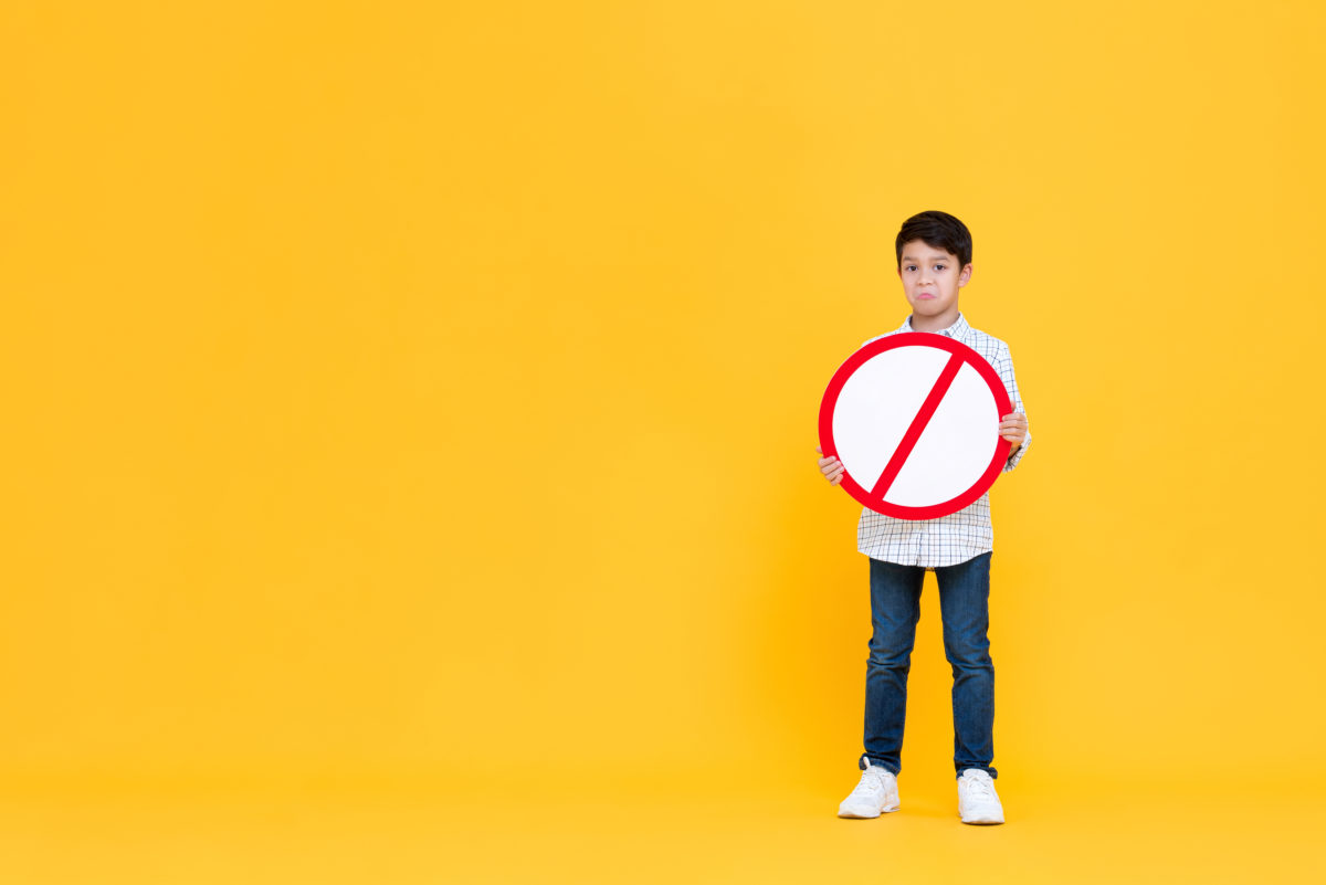 Portait of standing pouting young Asian boy holding red ban signage in yellow isolated studio background with copy space