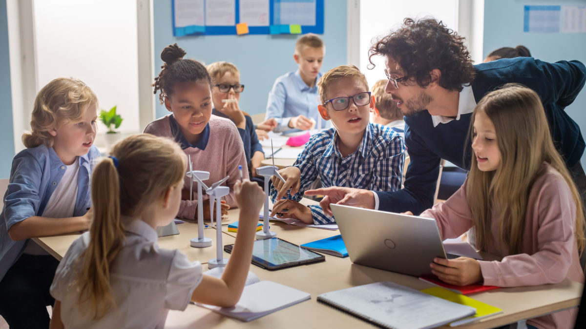 Elementary School Classroom: Enthusiastic Teacher Holding Tablet Computer Explains to a Brilliant Young Children How Wind Turbines Work. Kids Learning about Eco-Friendly Forms of Renewable Energy