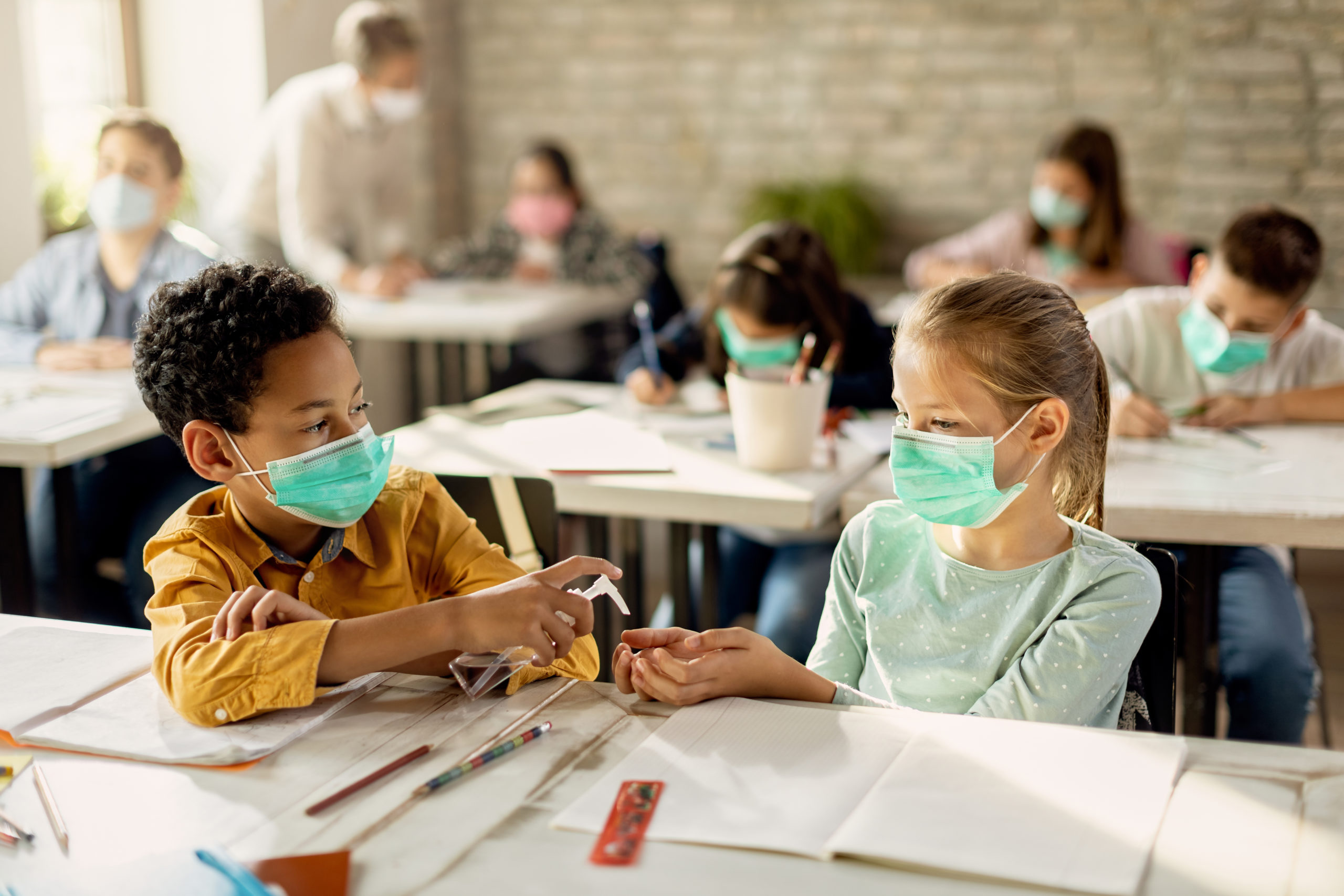 Two classmates with face masks sharing hand sanitizer in the classroom.