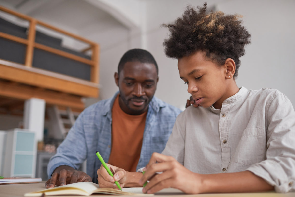 Portrait of teenage African-American boy doing homework or studying at home while sitting at desk with father helping him