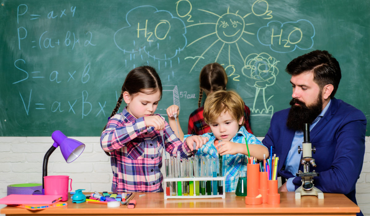 School club education. Chemistry themed club. Discover and explore properties of substances together. Interests and topic club. Teacher and pupils test tubes in classroom. Older kids help younger