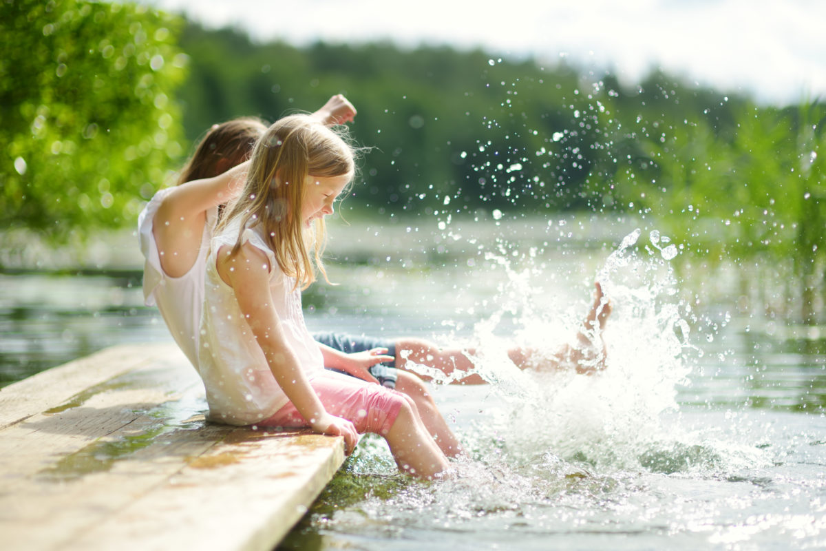 Two cute little girls sitting on a wooden platform by the river or lake dipping their feet in the water on warm summer day