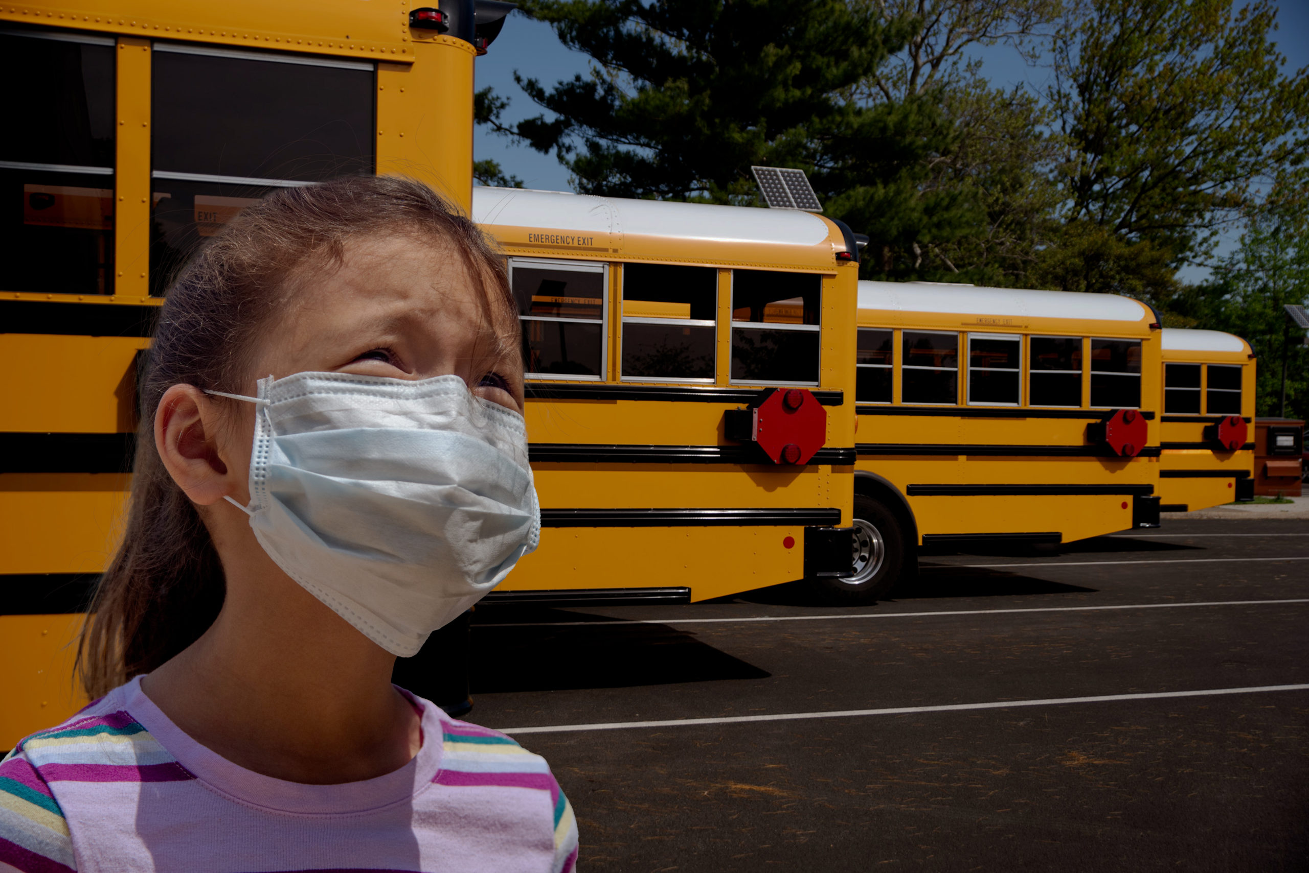 Young girl with concerned look in eyes stands in front of row of school buses wearing a mask to protect her from pandemic while riding Public transportation