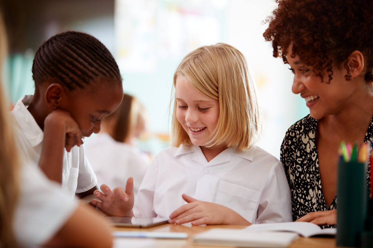 Female Teacher With Two Elementary School Pupils Wearing Uniform Using Digital Tablet At Desk