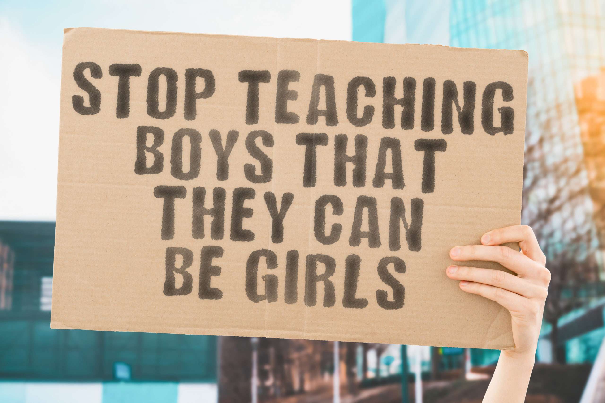 The phrase " Stop teaching boys that they can be girls " on a banner in hand with blurred background. Gay. Gays. LGBT. Education. Study. School. Same-sex. Couple