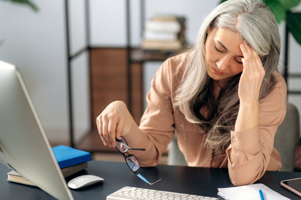 Tired upset mature gray-haired Asian, business woman, manager or teacher, takes a break from online work, massages her head with her hand, has a headache, sits at the table, took off glasses,need rest