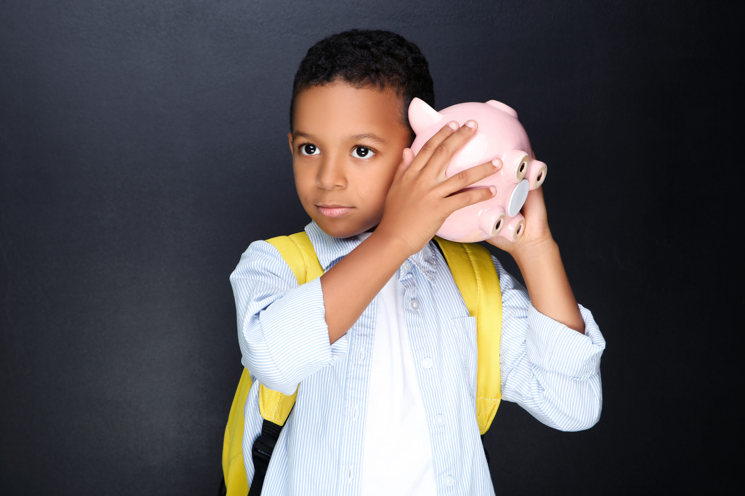 Young African American school boy with piggybank and backpack on black background