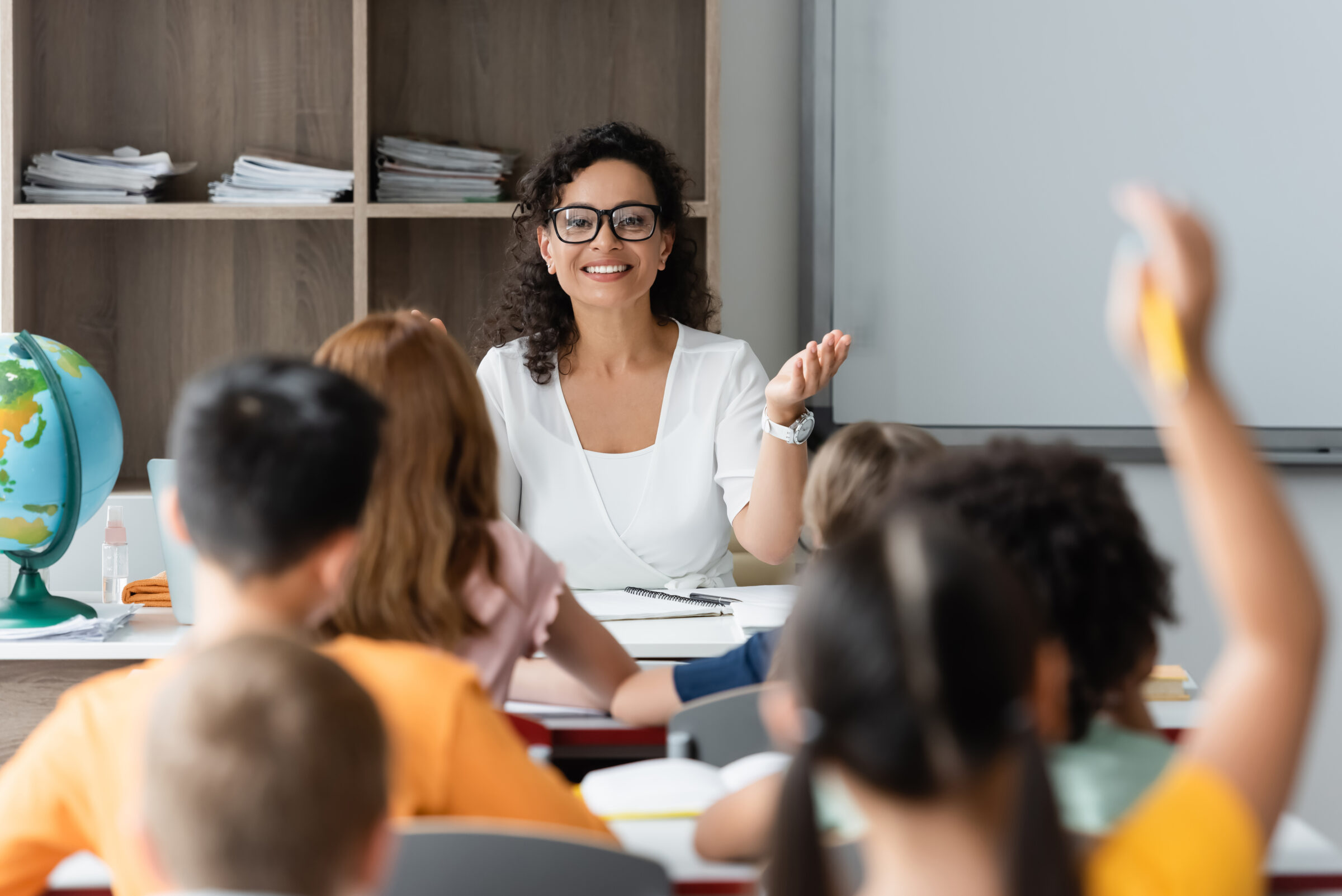 positive african american teacher pointing at blurred girl raising hand in classroom